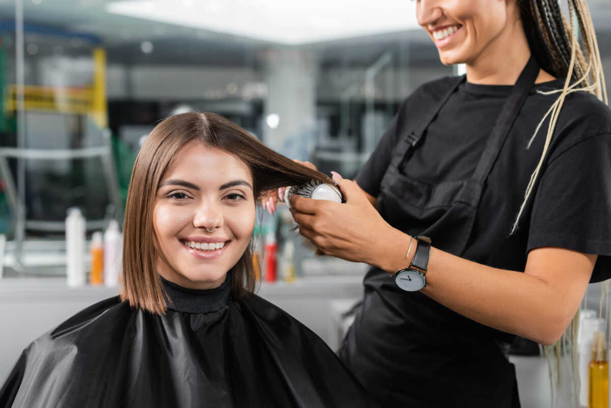 salon services, hairdresser with round brush styling hair of female customer, happy brunette woman