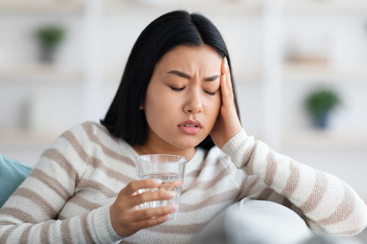 Young asian woman suffering from headache or migraine at home, holding glass of water and touching head temples after taking pill, beautiful korean lady feeling unwell, having acute pain, closeup