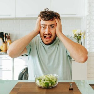 Sitting in kitchen and hold head with his hands, a young man looks desperately into the camera. Tired of eating lettuce and leafy greens. Healthy food and favorite cuisine. Healthy food concept.