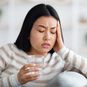 Young asian woman suffering from headache or migraine at home, holding glass of water and touching head temples after taking pill, beautiful korean lady feeling unwell, having acute pain, closeup